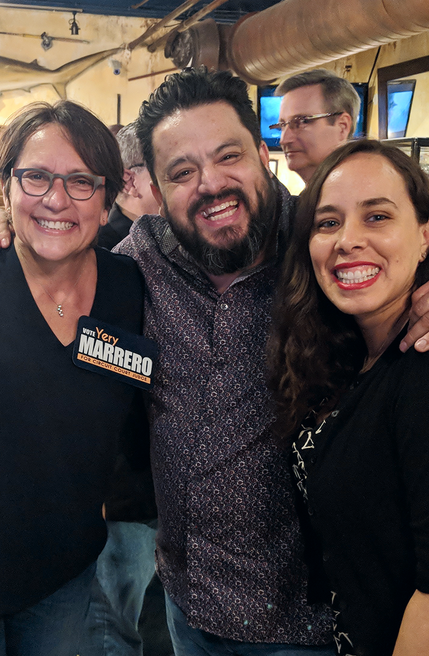 Group of three smiling people at a social event, wearing badges and casual attire.